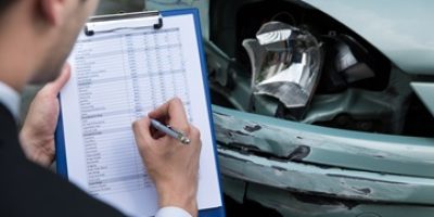 Side view of writing on clipboard while insurance agent examining car after accident